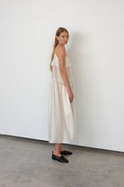 A woman stands in profile wearing a long, loose white sleeveless dress with dark brown flat shoes, set against a minimalist white wall and gray concrete floor.